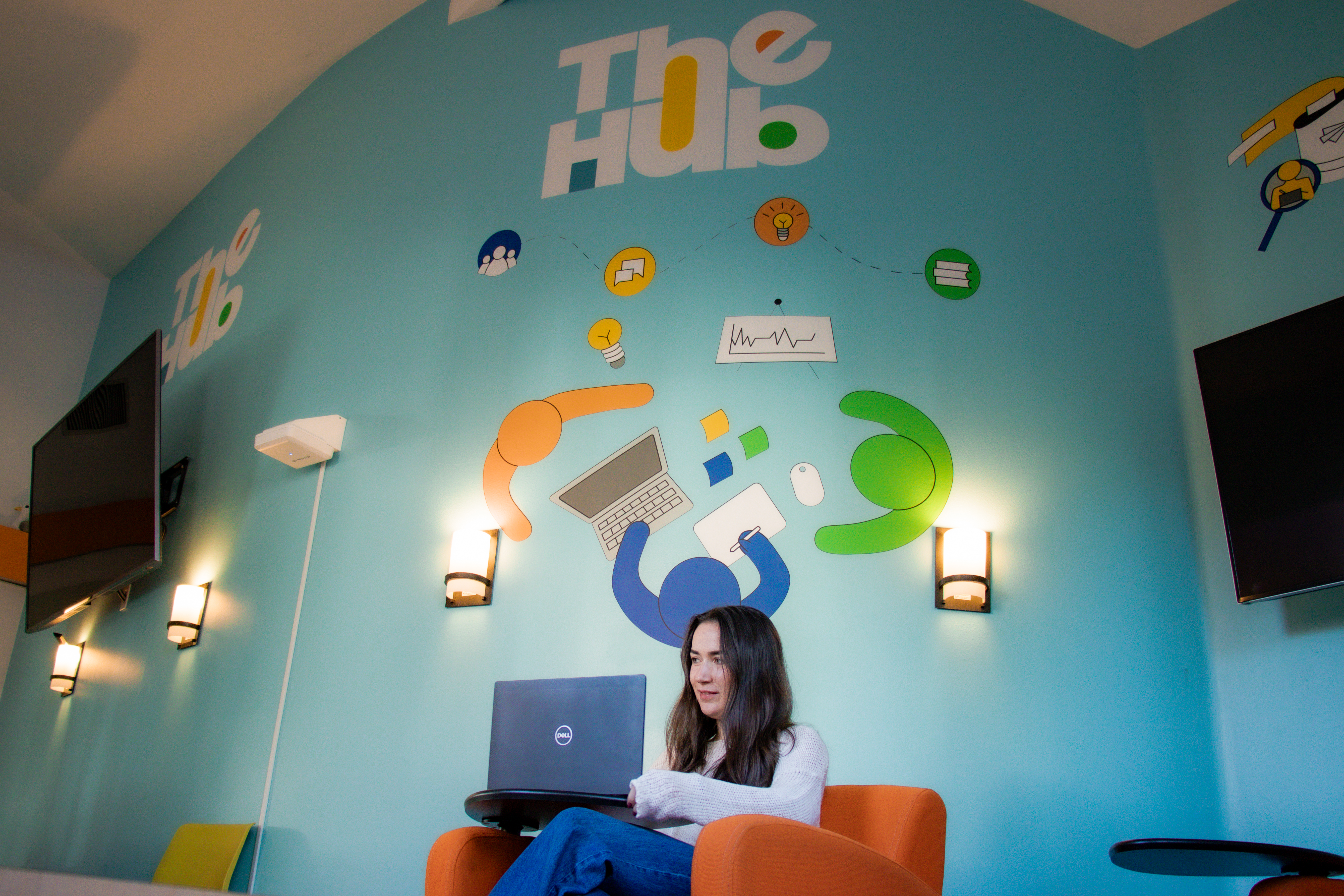 student working on a laptop in front of a wall with monitors and fun vinyl graphics of people